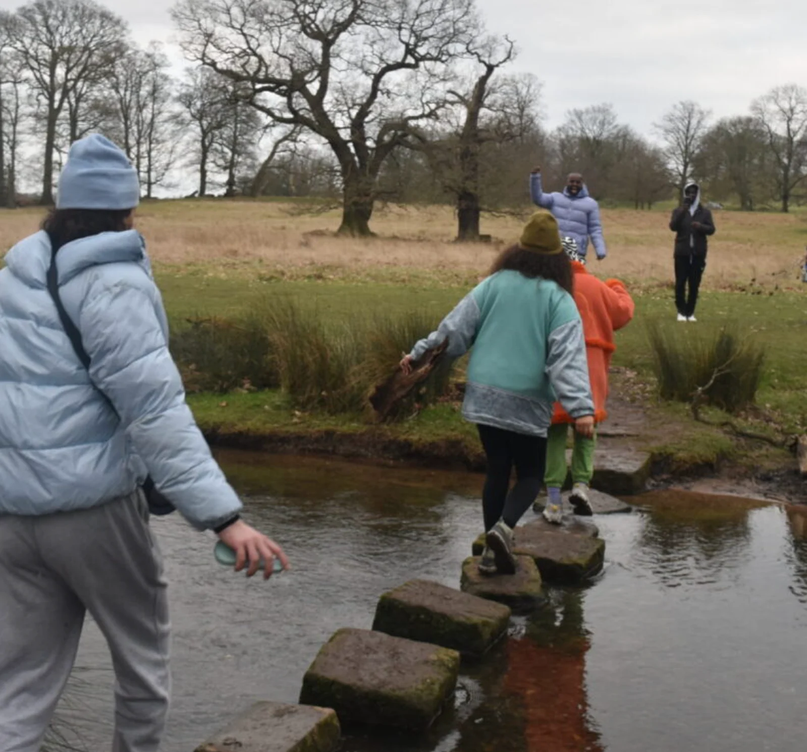 A group of people dressed for cold weather, crossing a stream using stepping stones. Two people wait on the bank